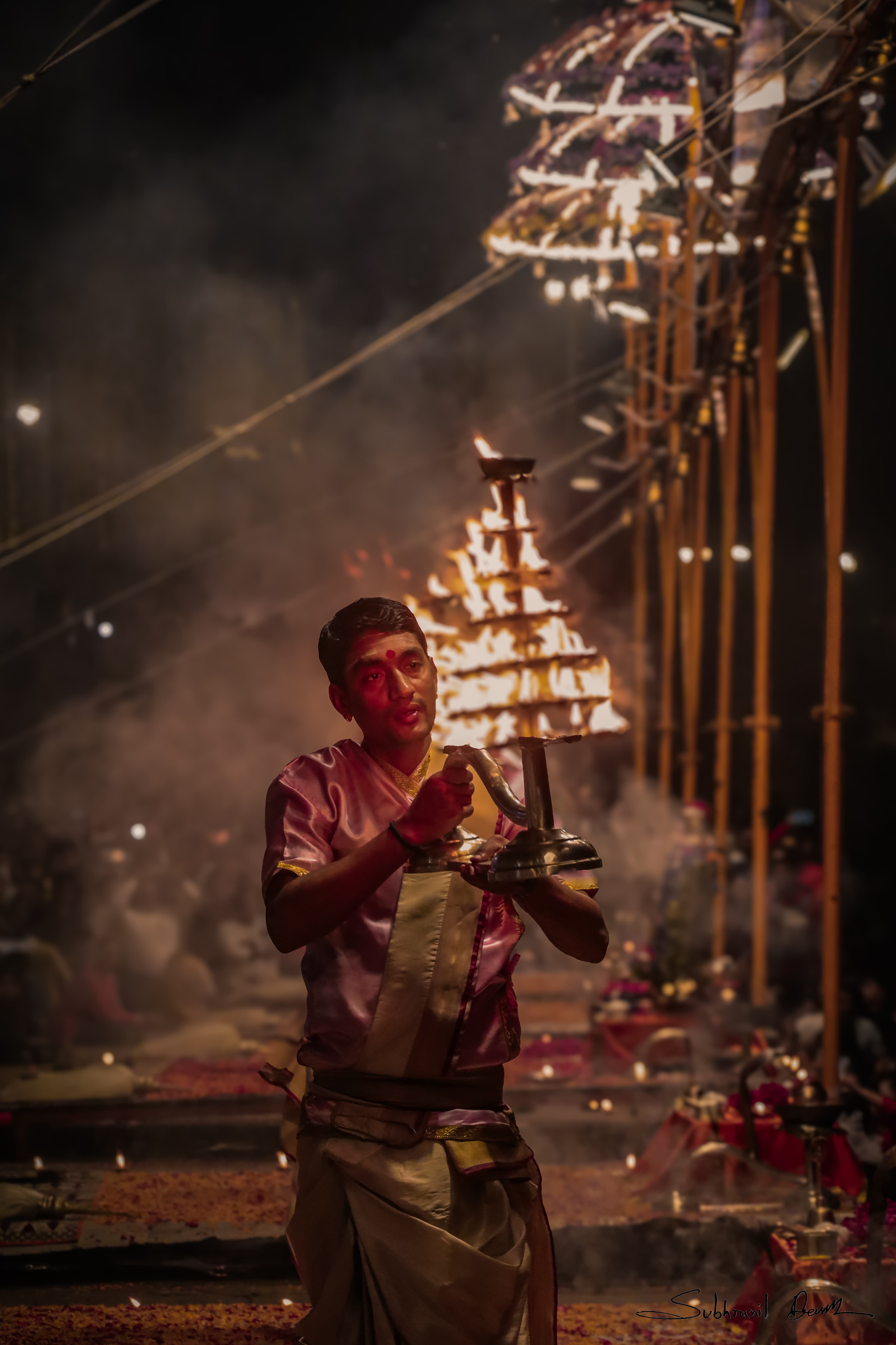 Amidst the sacred ghats of Varanasi, a priest performs the revered Ganga Aarti, holding a multi-tiered brass lamp as flames rise toward the heavens. The air is thick with smoke, chants, and the rhythmic sounds of bells, creating a mesmerizing spiritual atmosphere. This ritual, performed every evening, symbolizes the eternal bond between the Ganges and the devotees, illuminating the ancient city with divine energy and unwavering faith.