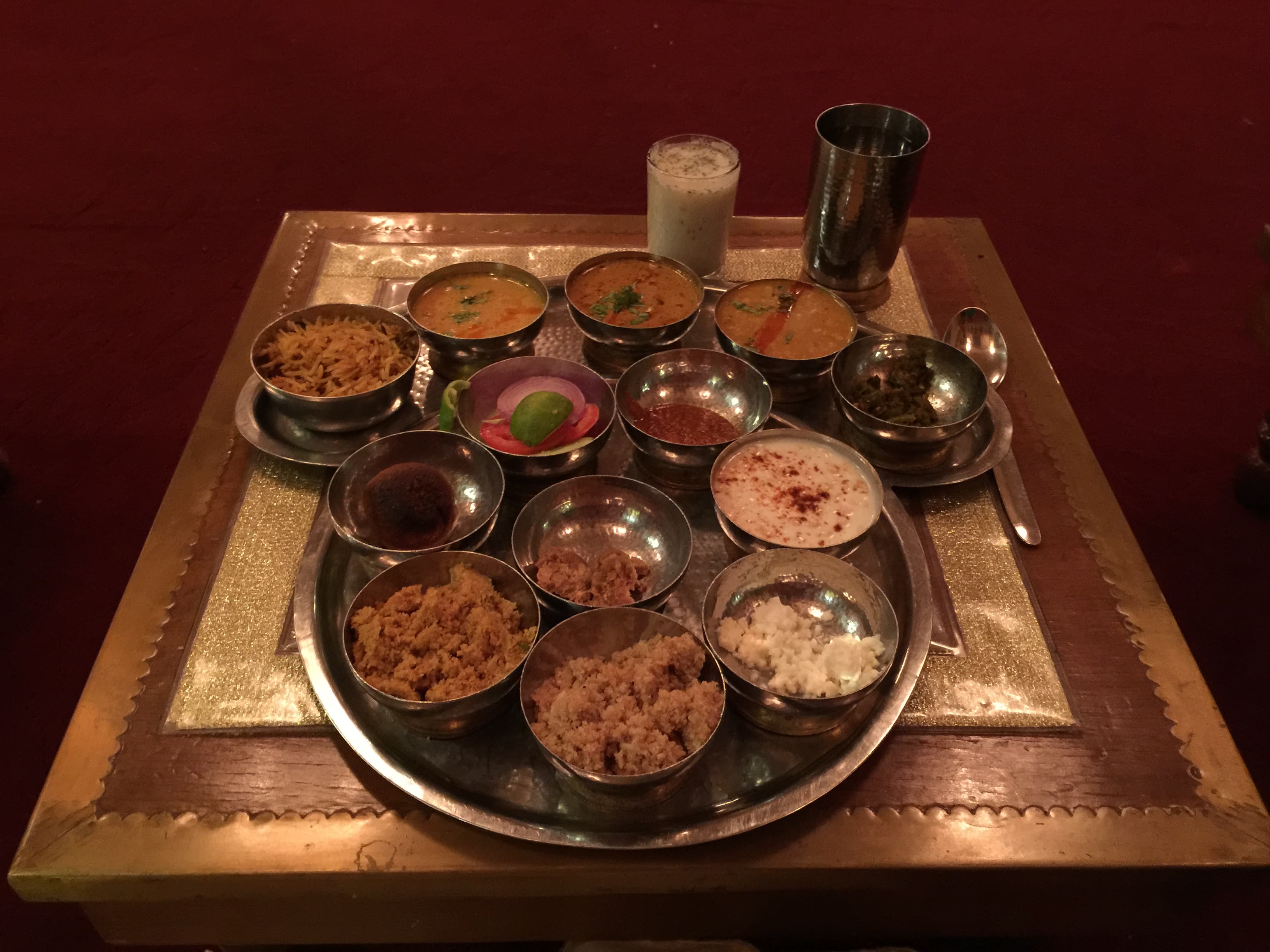 This is a traditional thali from the western state of Rajasthan, the land of the brave Rajputs. The thali features an essential Rajasthani dish called daal-bati-churma, which consists of baked batis served with delicious daal and sweet churma to delight the palate. In the center, a little jaggery is included to balance the flavors. There are two subzis: one made with aloo-pyaz and the other with mixed vegetables, accompanied by various types of rotis—wheat, bajra, jowar, etc.—and a side of flavorful red garlic chutney. Accompaniments include homemade churned butter, salad, raita, and the famous Rajasthani chaas. A serving of rice is presented in the form of pulav, slightly sweeter, with some mirchi pickle to tantalize the taste buds. This complete meal, enhanced by local dancers, is an experience that cannot be missed.